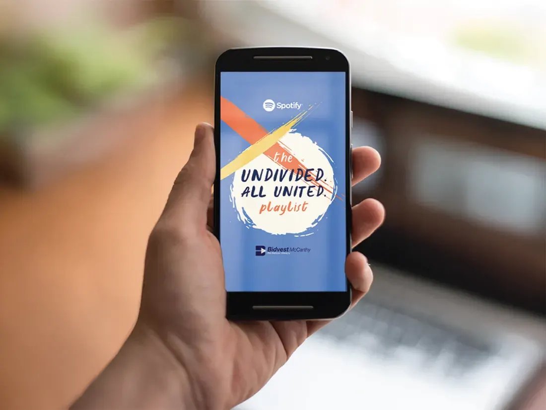 Hand holding a phone showing the Undivided. All United. playlist cover: brushstroke mark in orange and yellow on blue.